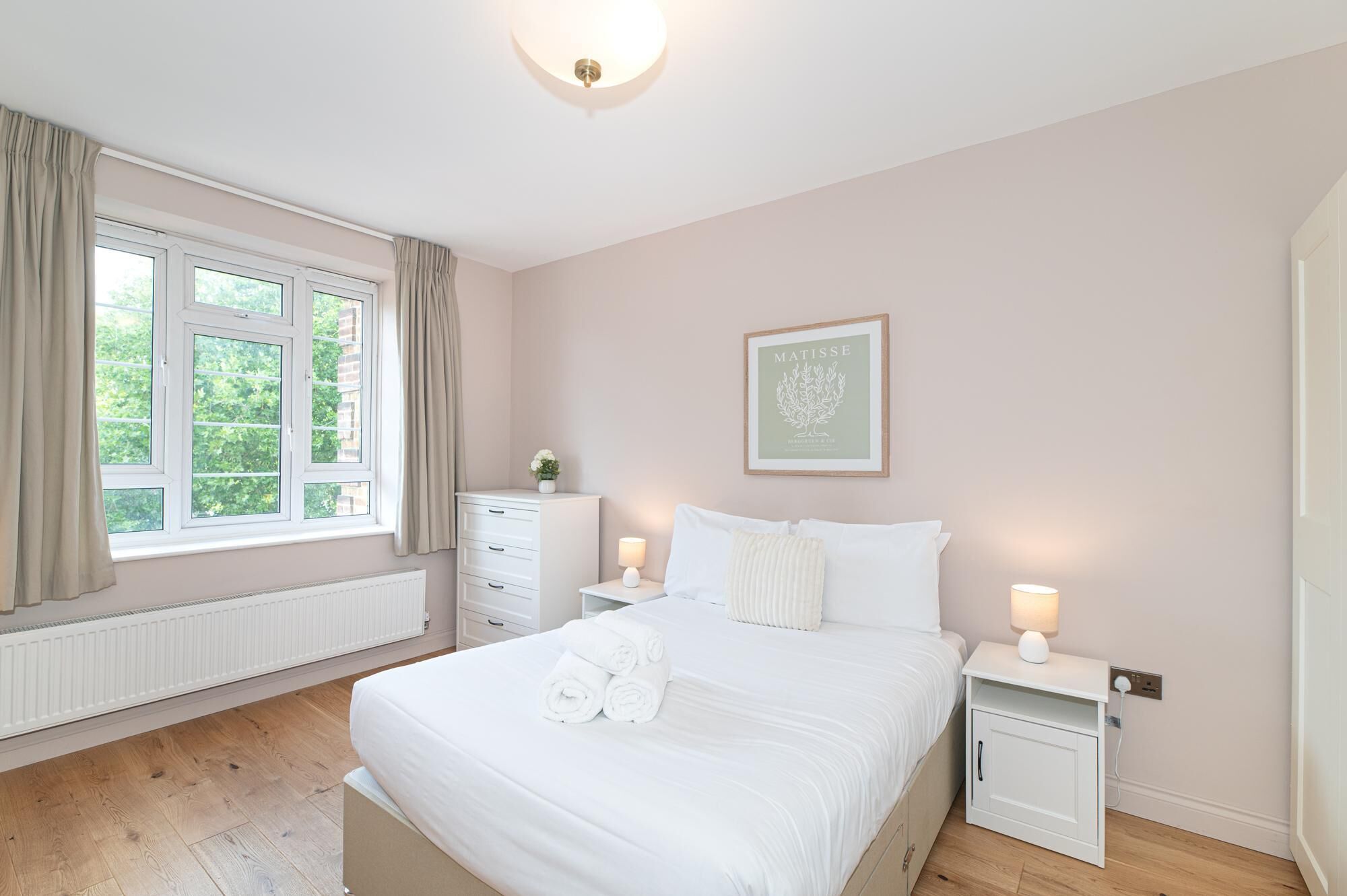 Quarto com cama, mesas de cabeceira, radiador, janela com cortinas e um quadro na parede.