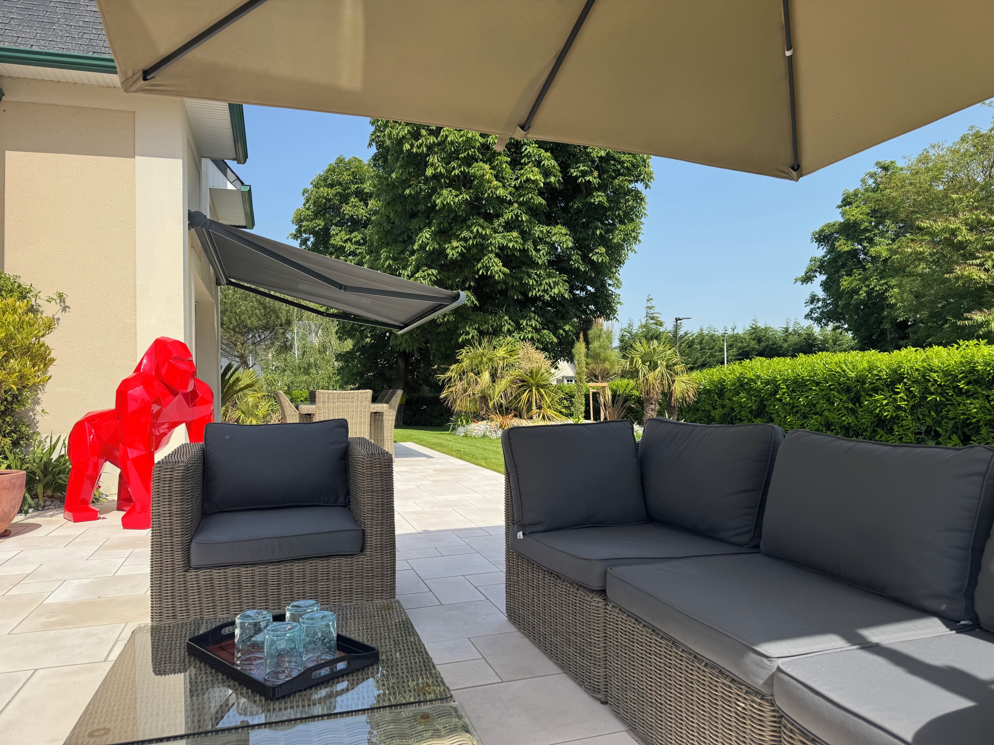 Een buitenzitgedeelte met rieten meubels en een glazen tafel, onder een grote parasol.