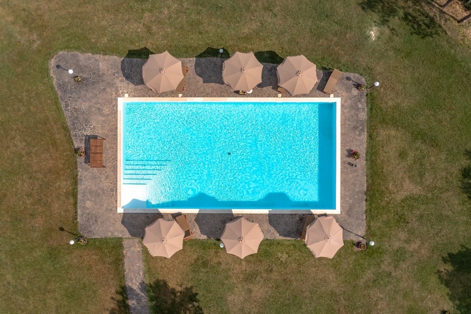 Vista aérea de una piscina rectangular rodeada de baldosas de piedra, con múltiples sombrillas y una terraza de madera.