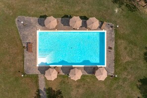 An aerial view of a rectangular swimming pool surrounded by stone tiles, with multiple umbrellas and a wooden deck.