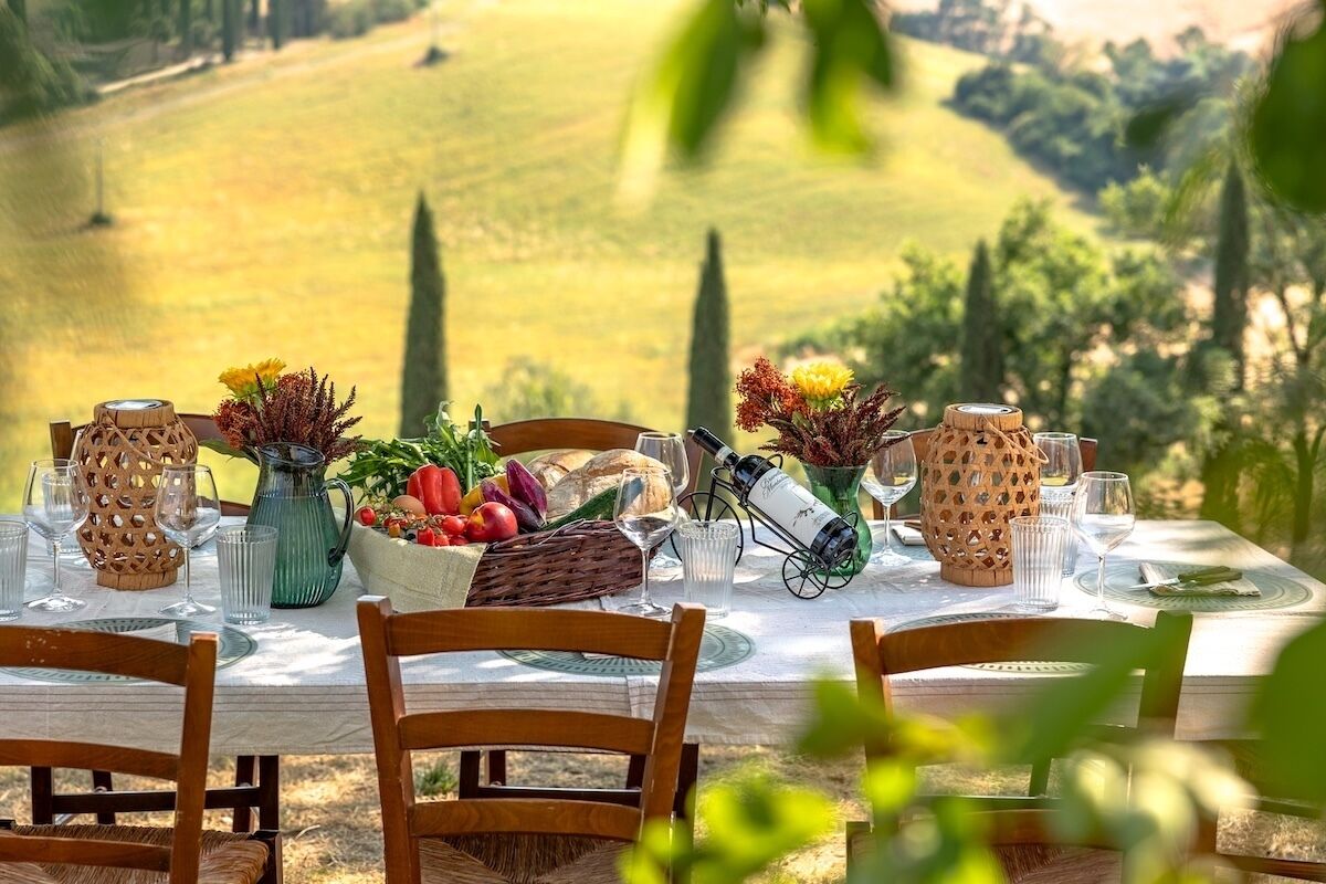 A table set for a meal with a scenic countryside view, including a basket of fruits and a wine bottle.
