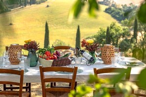 Une table dressée pour un repas, avec une vue pittoresque sur la campagne, comprenant un panier de fruits et une bouteille de vin.