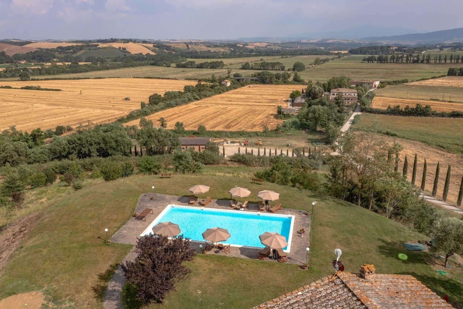 A pool area with lounge chairs and umbrellas, surrounded by a rural landscape with fields and trees.