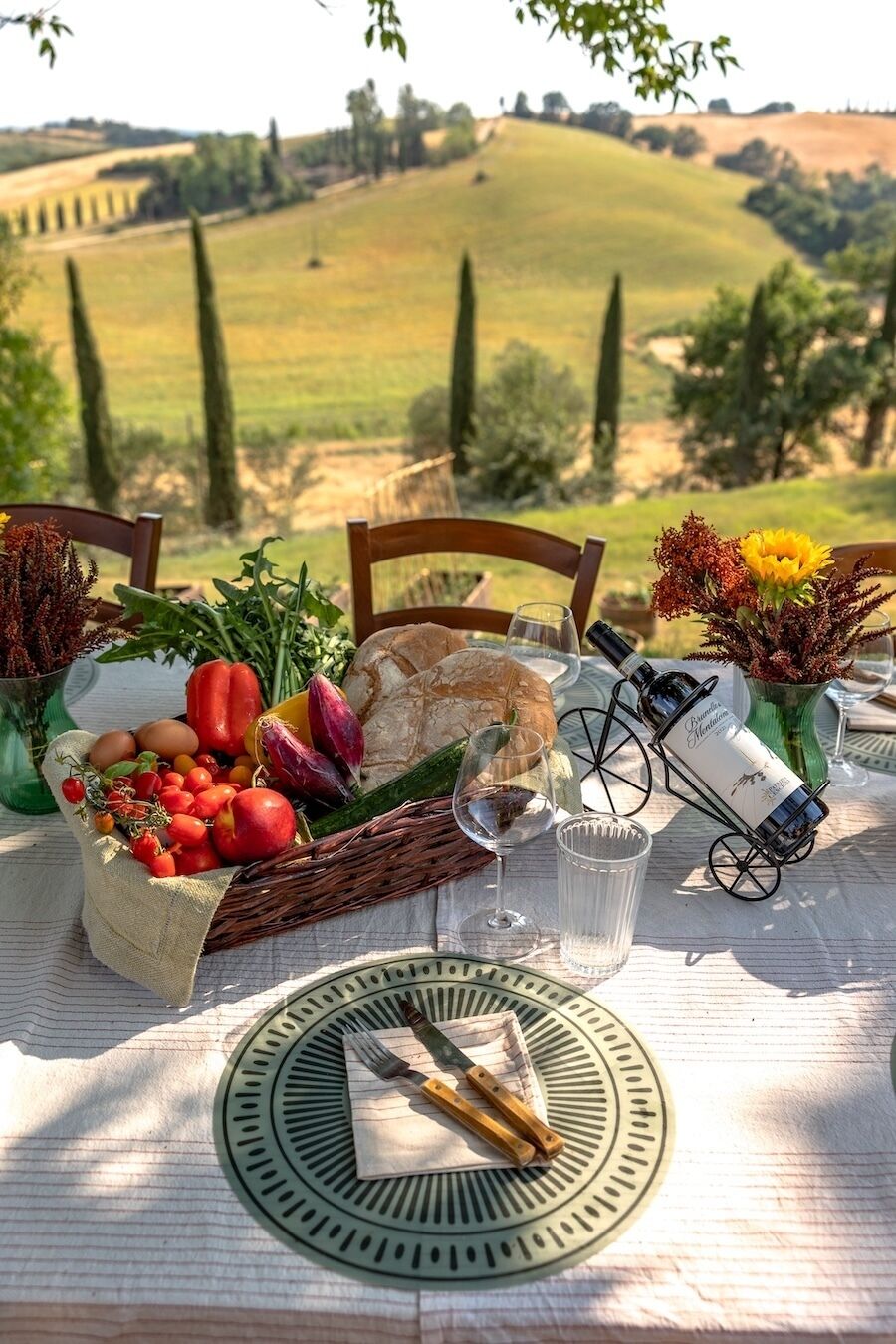 Mesa al aire libre con vista a colinas ondulantes, una cesta de verduras frescas, una botella de vino y una decoración rústica.