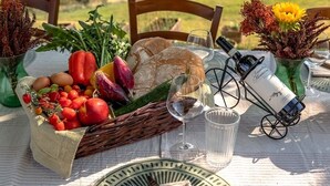 Tavolo apparecchiato all'aperto con vista su dolci colline, un cesto di verdure fresche, una bottiglia di vino e un'atmosfera rustica.