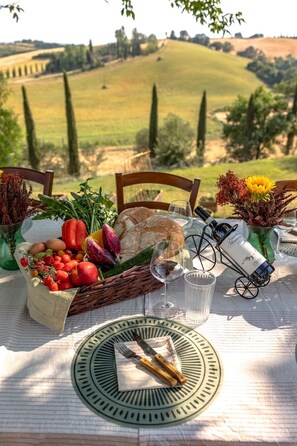 A table set outdoors with a view of rolling hills, a basket of fresh vegetables, a bottle of wine, and a rustic table setting. - Villa Comunello (Monteroni d'Arbia SI)