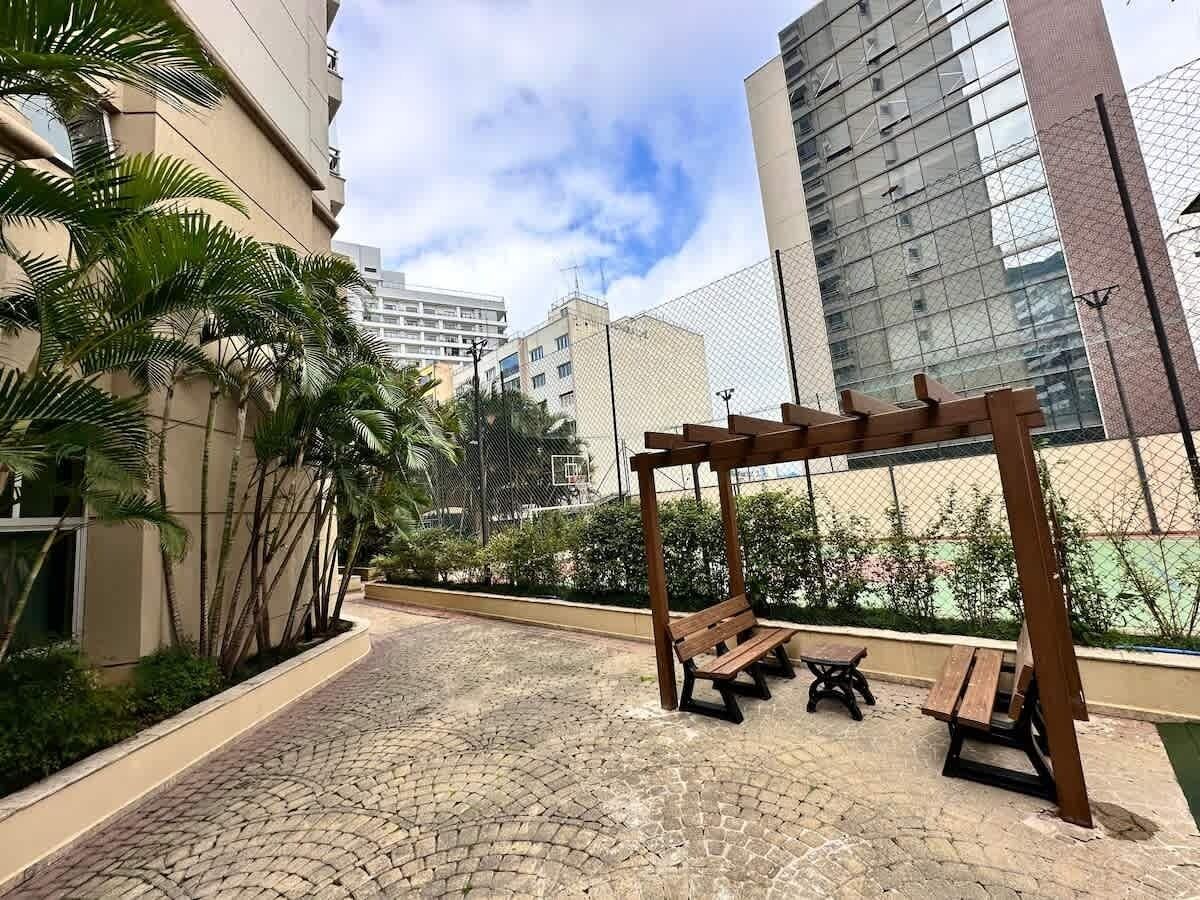 Um espaço ao ar livre pavimentado com um banco de madeira e uma pequena mesa, rodeado por vegetação e um cercado.