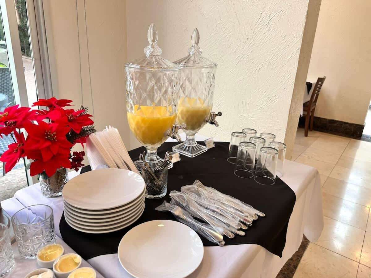 A table set with plates, cutlery, and two punch bowls with yellow contents, likely juice or punch, in a room with a tiled floor and a window with a view.