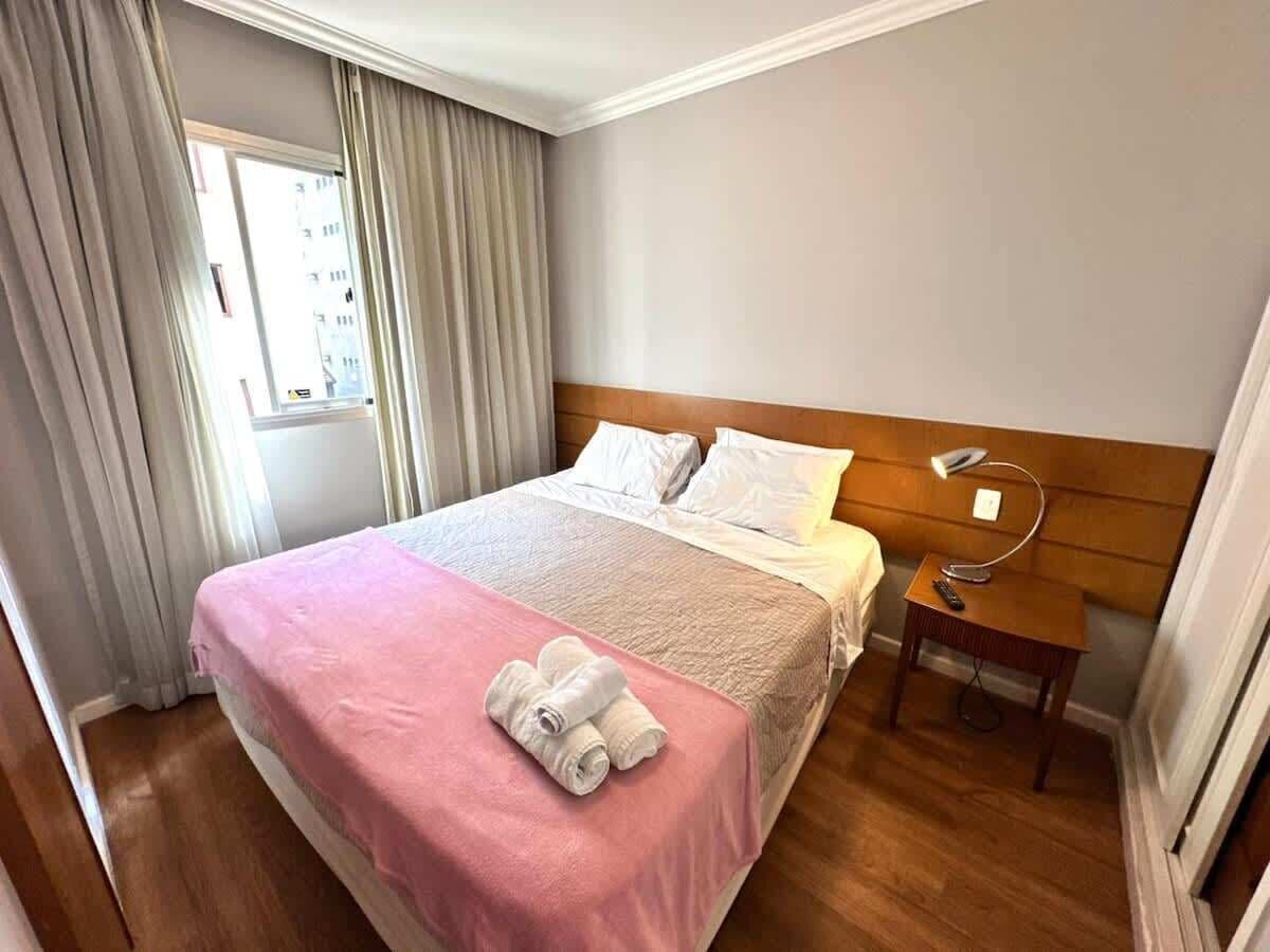 Een hotelkamer met een bed, nachtkastje, lamp en een raam met gordijnen.