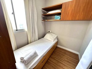 Ein kleines Zimmer mit einem Einzelbett, Holzboden, weißer Bettwäsche und einem Regal über dem Bett.