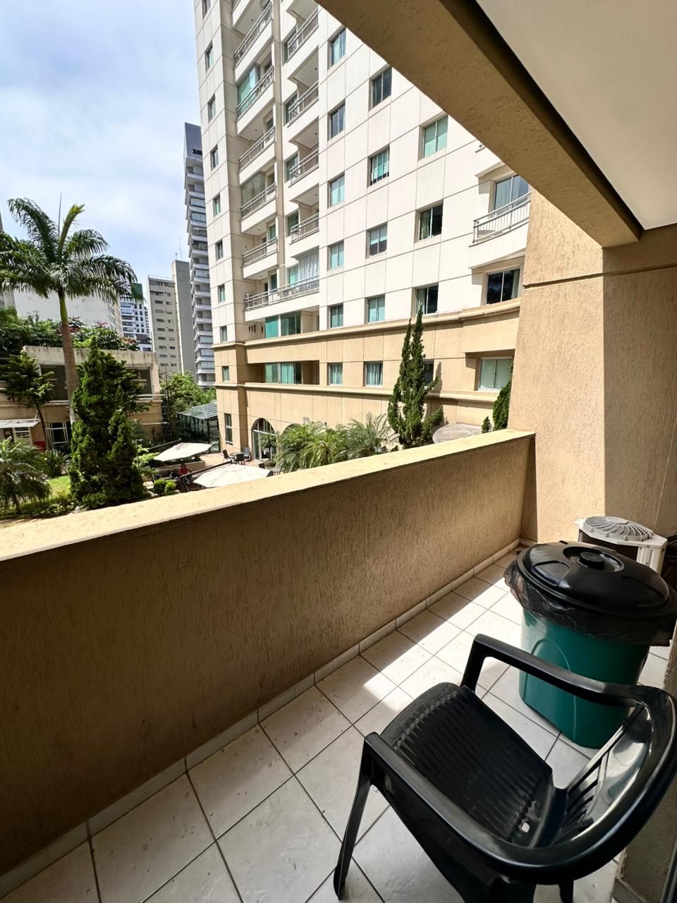 A balcony with a chair, a trash bin, and a view of a high-rise building with multiple floors and balconies.