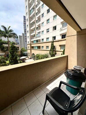 Un balcón con una silla, un basurero y vistas de un edificio de gran altura con varios pisos y balcones.