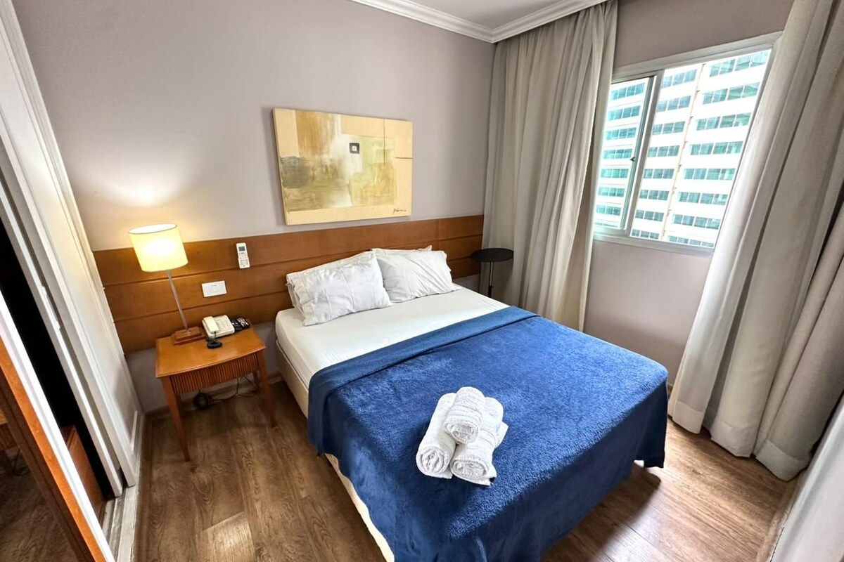 Ein Hotelzimmer mit einem Bett, einem Nachttisch mit Lampe, einem Bild an der Wand und einem Fenster mit Vorhängen.