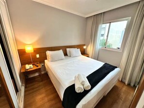 Habitación de hotel con cama, mesita de noche, lámpara y ventana con cortinas.