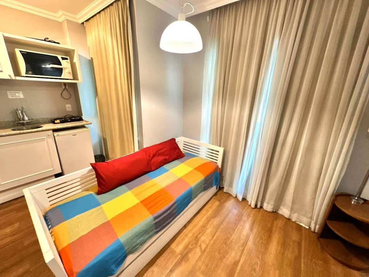 Ein Einzelbett mit farbenfroher Decke, ein weißer Nachttisch, eine Mikrowelle und eine kleine Küchenzeile.