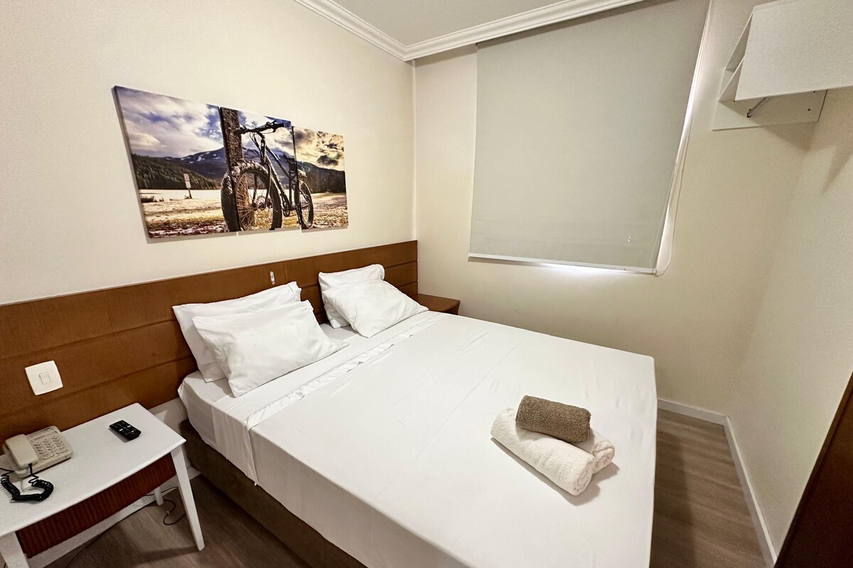 Ein ordentlich bezogenes Bett mit weißen Leinentüchern, ein Nachttisch mit Telefon und Fernbedienung sowie ein Gemälde einer Berglandschaft an der Wand.