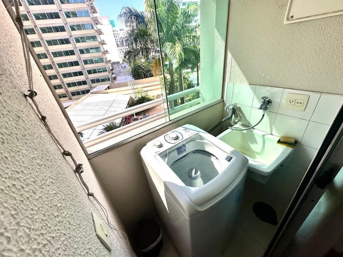 Uma lavanderia compacta com máquina de lavar, pia e janela com vista para um edifício e palmeiras.