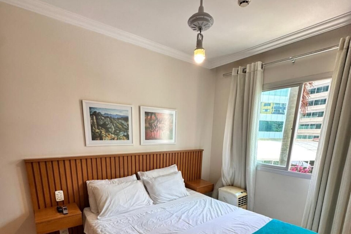 Una habitación de hotel con cama, mesita de noche, ventilador de techo y ventana con cortinas.