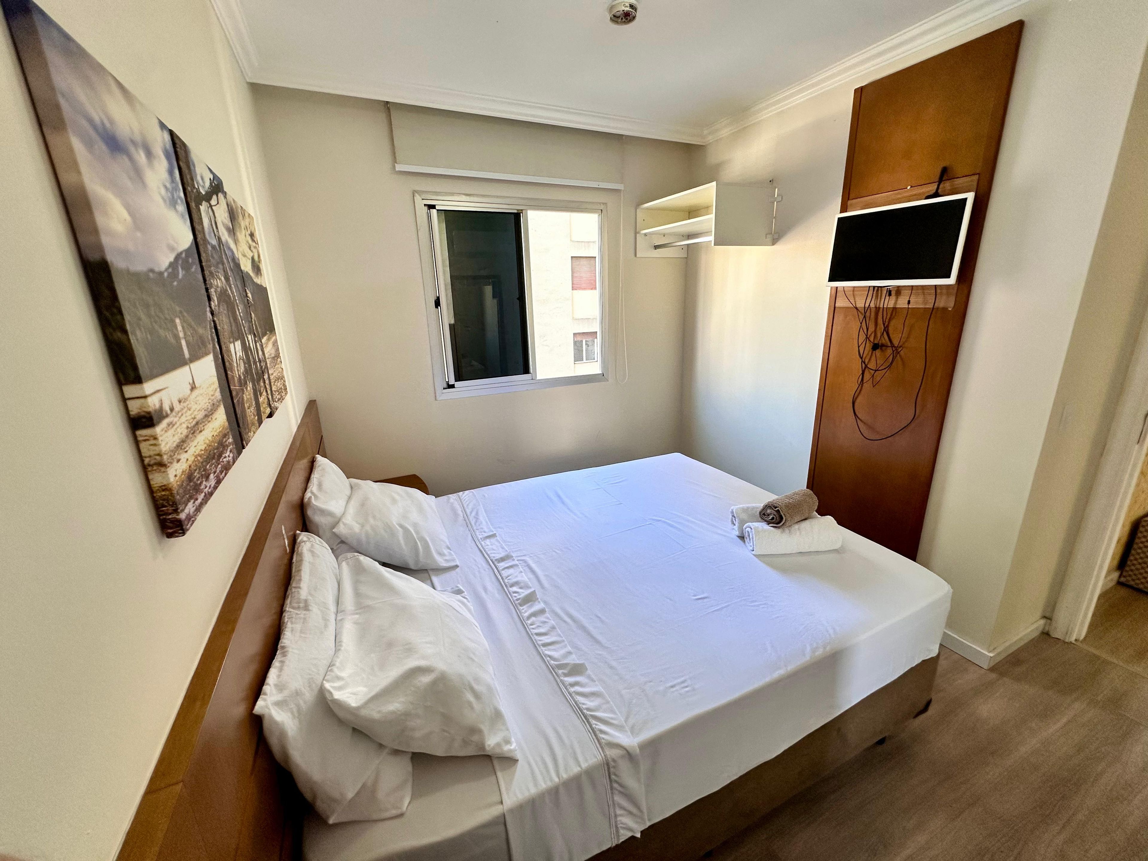 Un dormitorio con cama, ventana, televisor montado en la pared y puerta.