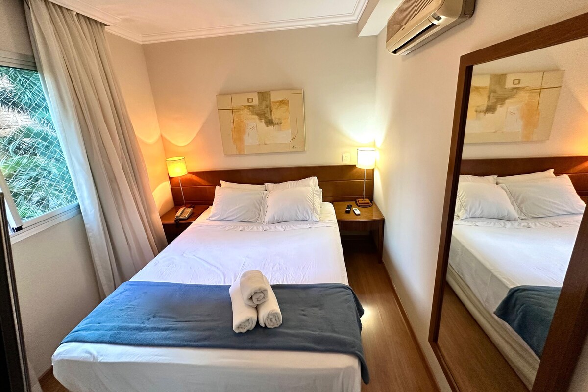 Ein Hotelzimmer mit zwei Betten, einem großen Spiegel, einer Wand-Klimaanlage und einem Fenster mit Vorhängen.