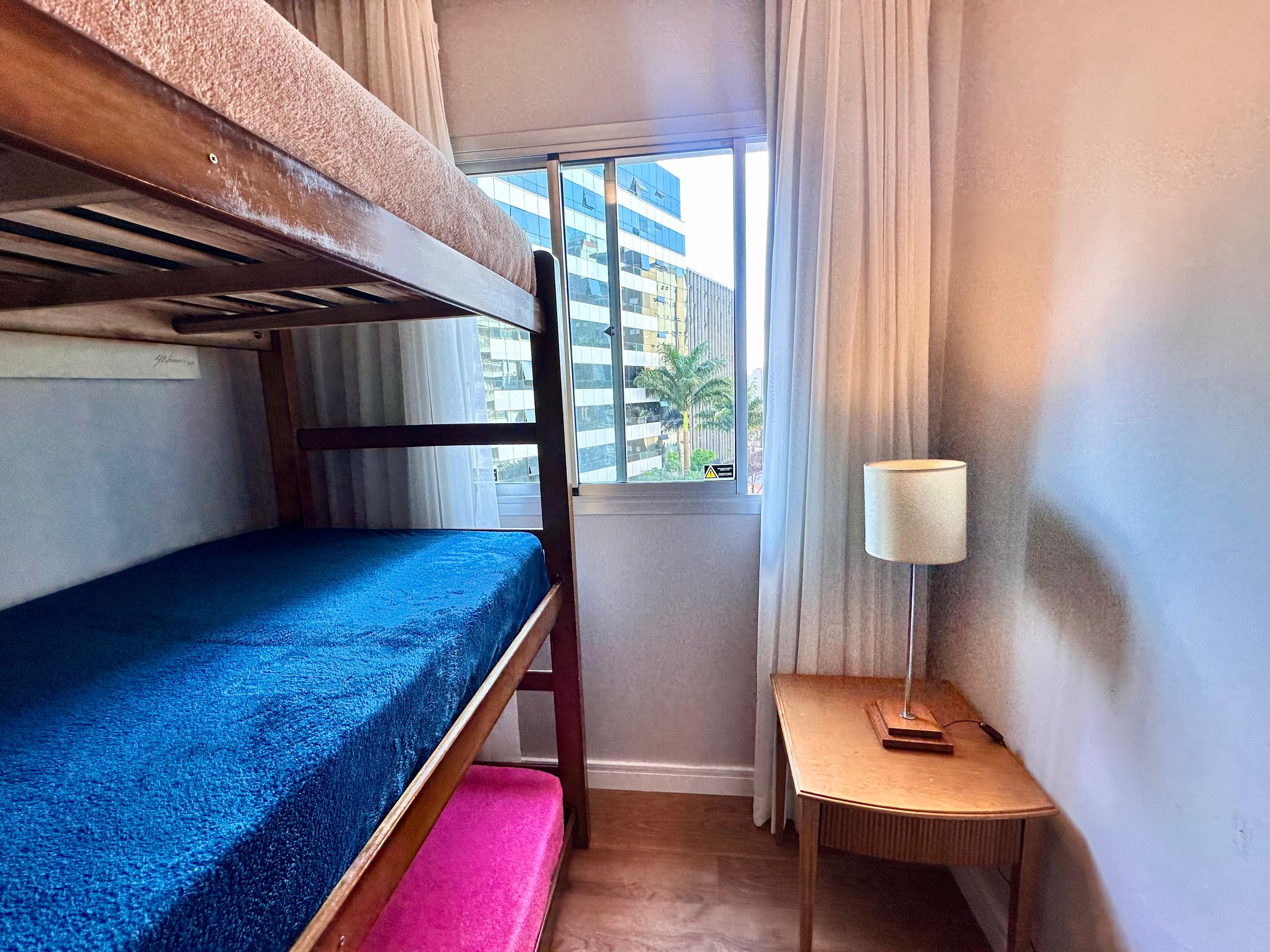 Une petite chambre avec un lit superposé, une petite table avec une lampe et une fenêtre donnant sur des immeubles.