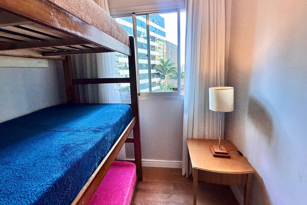 Ein kleines Zimmer mit einem Etagenbett, einem kleinen Tisch mit Lampe und einem Fenster mit Blick auf Gebäude.