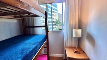 Une petite chambre avec un lit superposé, une petite table avec une lampe et une fenêtre donnant sur des immeubles.