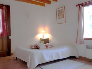 A single bed with a white bedspread and two pillows, a bedside lamp, a framed picture on the wall, and a window with curtains.