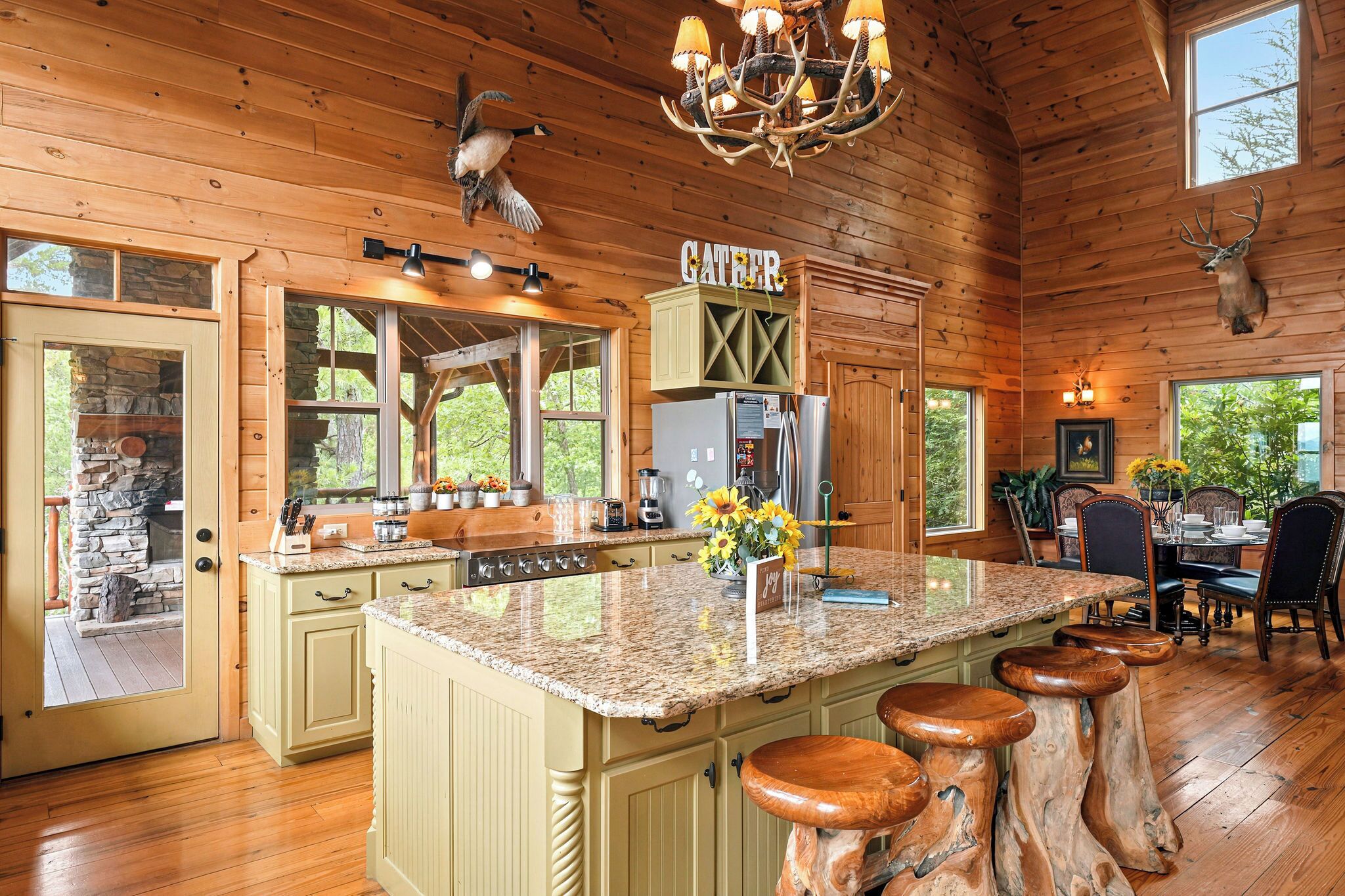 A cabin interior with a kitchen island, bar stools, a dining area, and a stone fireplace.