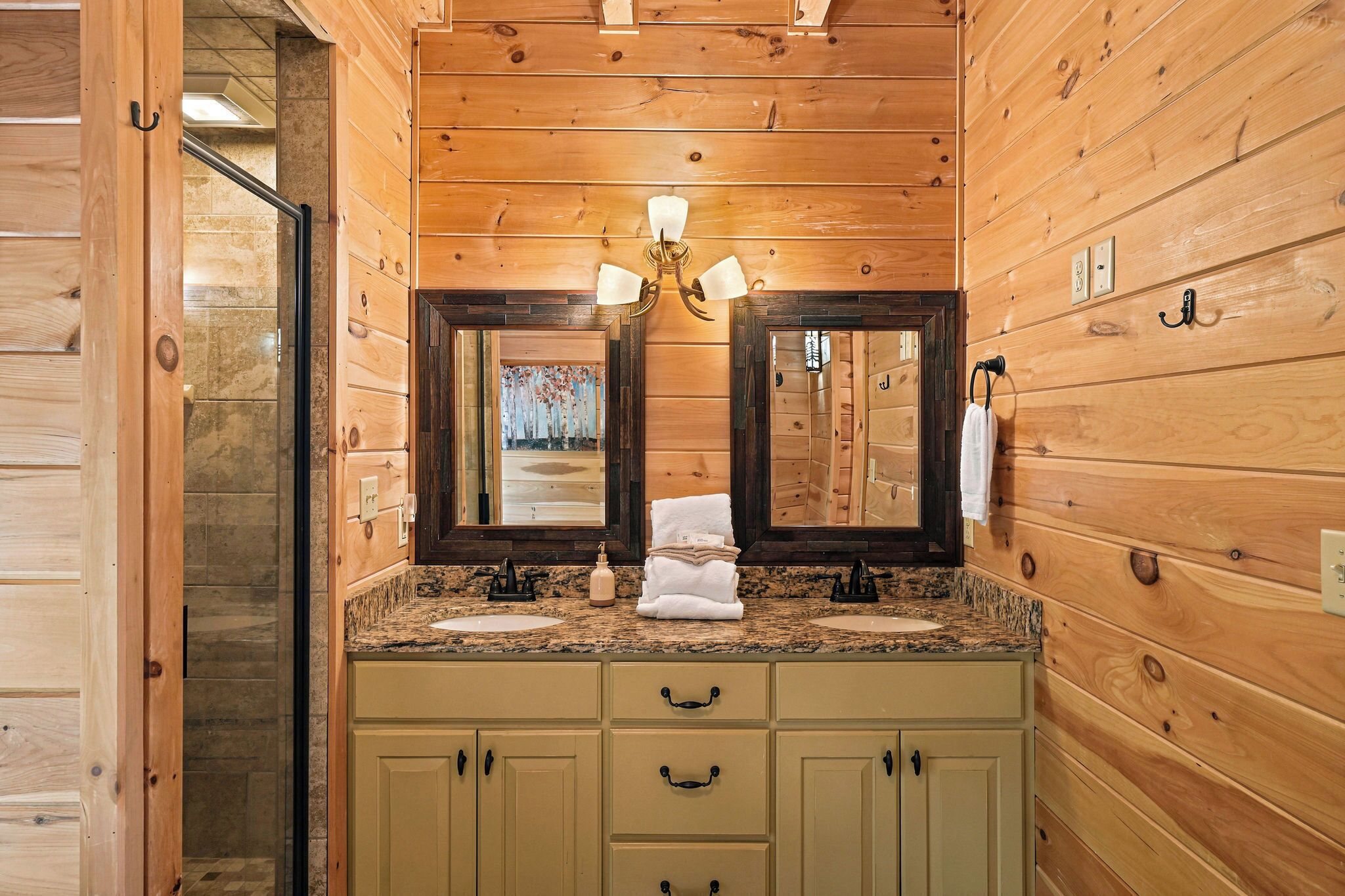 A wooden cabin bathroom with a double sink, two mirrors, a chandelier, and a towel rack.