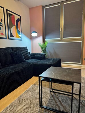 A modern living room with a sectional sofa, a small table, and a large window with blinds. - Apartment in Botanical Gardens, Old Trafford (Manchester)