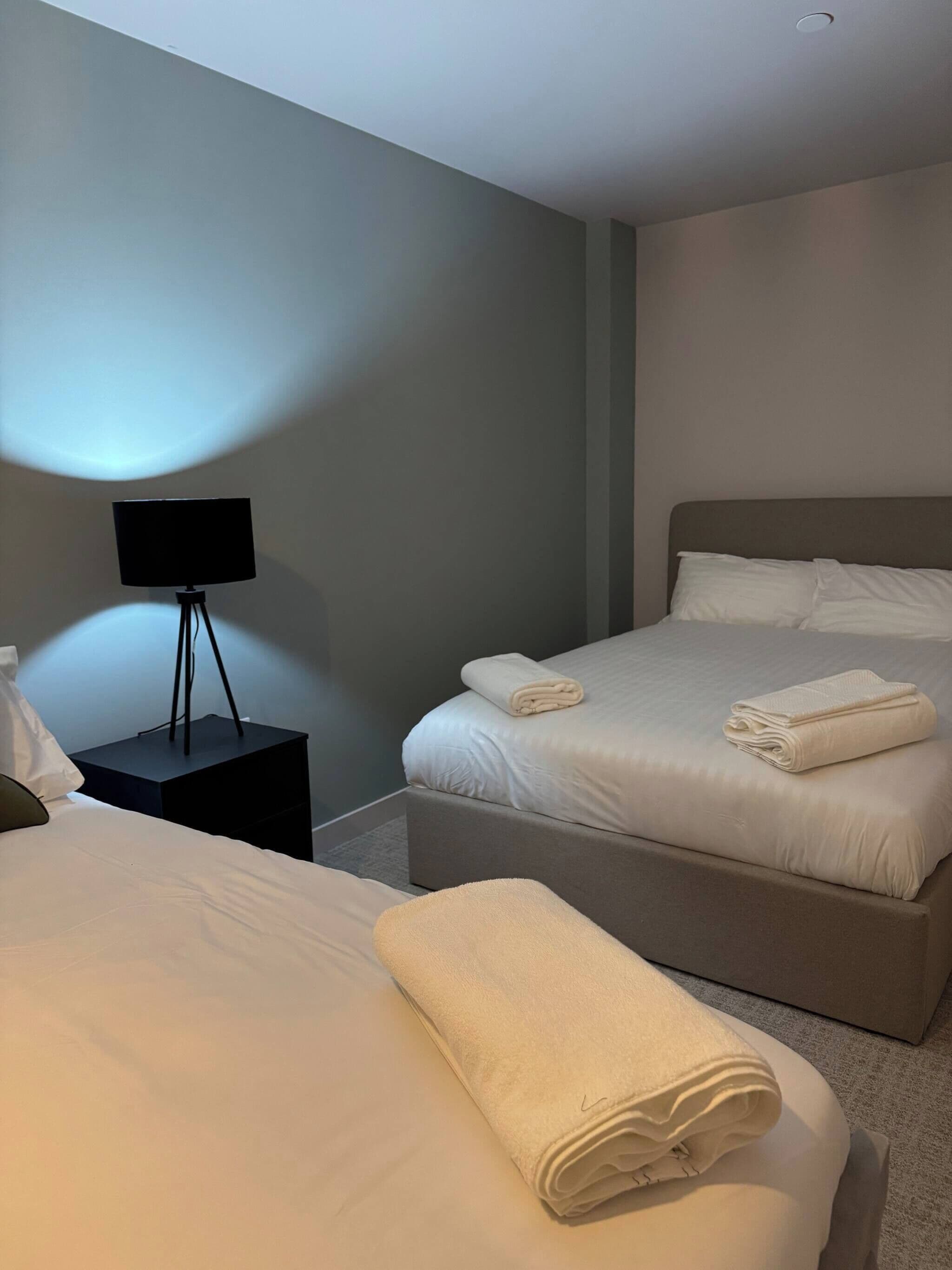 A hotel room with two beds, a nightstand with a lamp, and a wall-mounted light.
