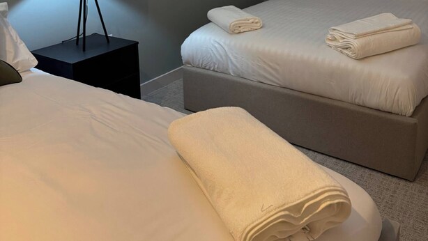 A hotel room with two beds, a nightstand with a lamp, and a wall-mounted light. - Apartment in Botanical Gardens, Old Trafford (Manchester)