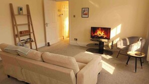 A living room with a beige sofa, a television displaying a fireplace, a small round table, and a chair. - 1BR Apartment in Chislehurst High Street Sleeps 3 (Chislehurst)