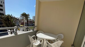 Uma varanda com mesa e cadeiras brancas, chão de azulejos e vista para edifícios e palmeiras.