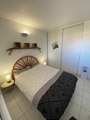 Ein Schlafzimmer mit hölzernem Kopfteil, einem Bett mit gemusterten Betttextilien, einem Nachttisch mit Lampe und einem Wandregal mit Dekorationen.