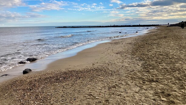 Uma praia com areal pedregoso, um cais distante e céu claro.