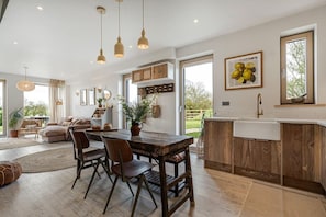 A modern kitchen with wooden cabinets, a central island, pendant lighting, and a large window providing natural light. - The Fold | A Tucked-Away 2 Bedroom Retreat In Buckinghamshire (Milton Keynes)