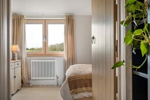 A bedroom with a bed, a radiator, a window with curtains, a bedside table with a lamp, and a plant. - The Fold | A Tucked-Away 2 Bedroom Retreat In Buckinghamshire (Milton Keynes)