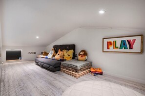 Un dormitorio acogedor en el ático, con techo inclinado, un ventanal grande, una cama con juguetes suaves, un cartel enmarcado que dice 'PLAY' y una pila de libros.
