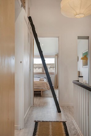 Un pasillo con una escalera negra apoyada en la pared, una cama en la habitación contigua y una lámpara en el techo.