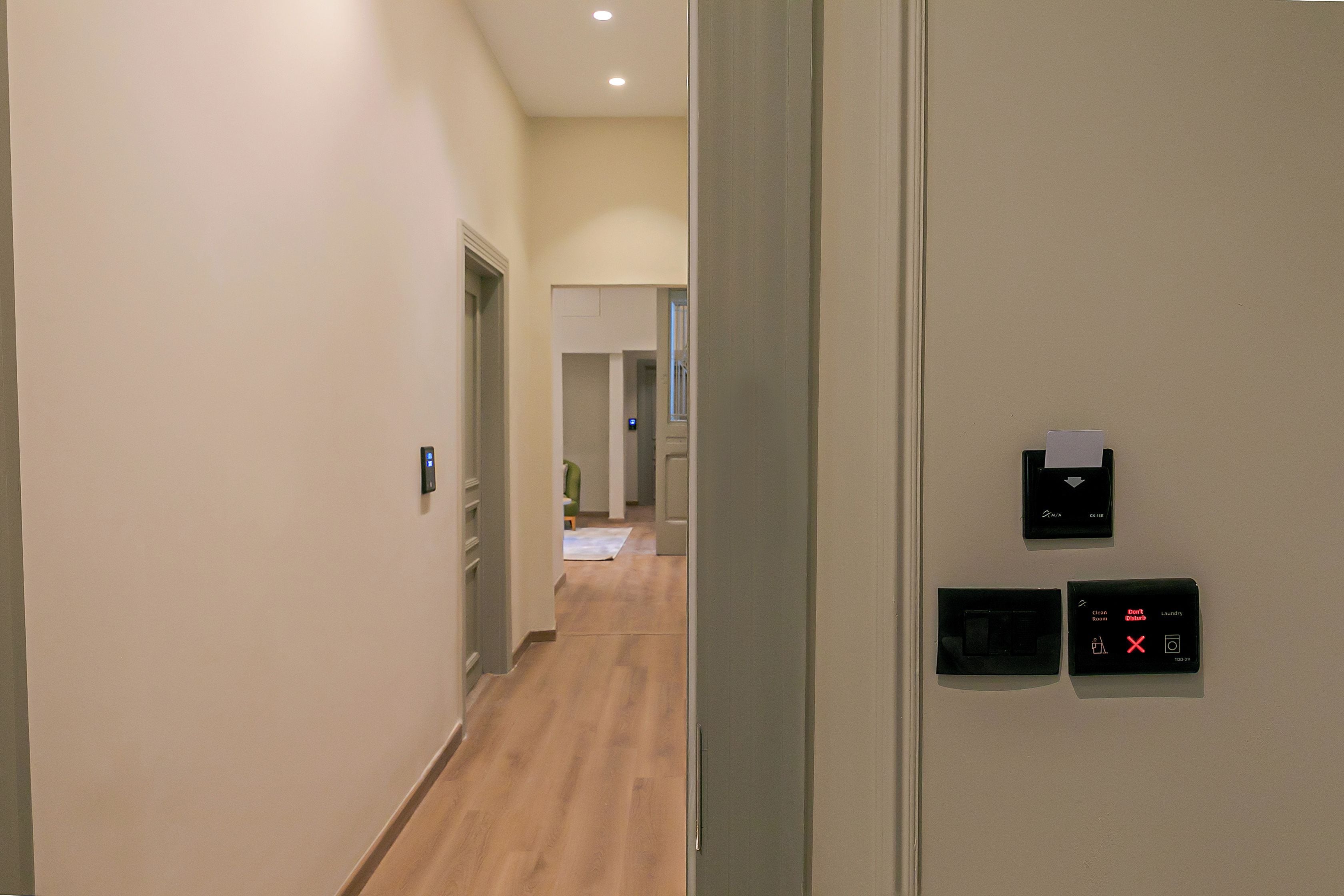 Un pasillo con suelo de madera, una puerta a la izquierda y un panel de control fijado a la pared con pantalla digital.