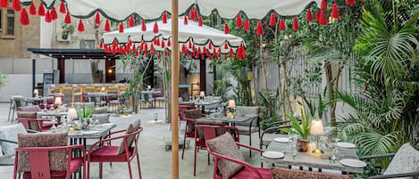 Un área de comedor al aire libre con mesas y sillas, un toldo blanco y flecos rojos.