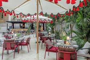 Un área de comedor al aire libre con mesas y sillas, un toldo blanco y flecos rojos.