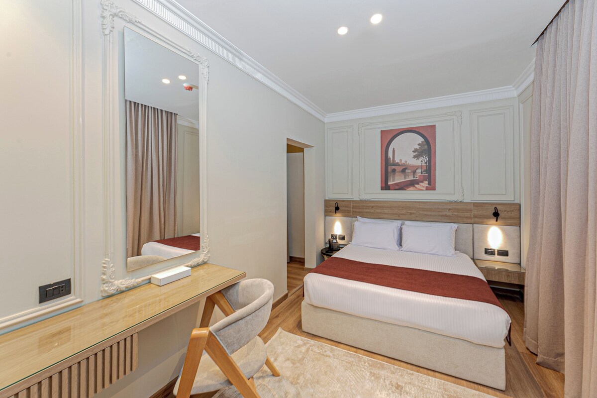 A hotel room with a large bed, a wooden desk, a chair, a mirror, and a painting on the wall.