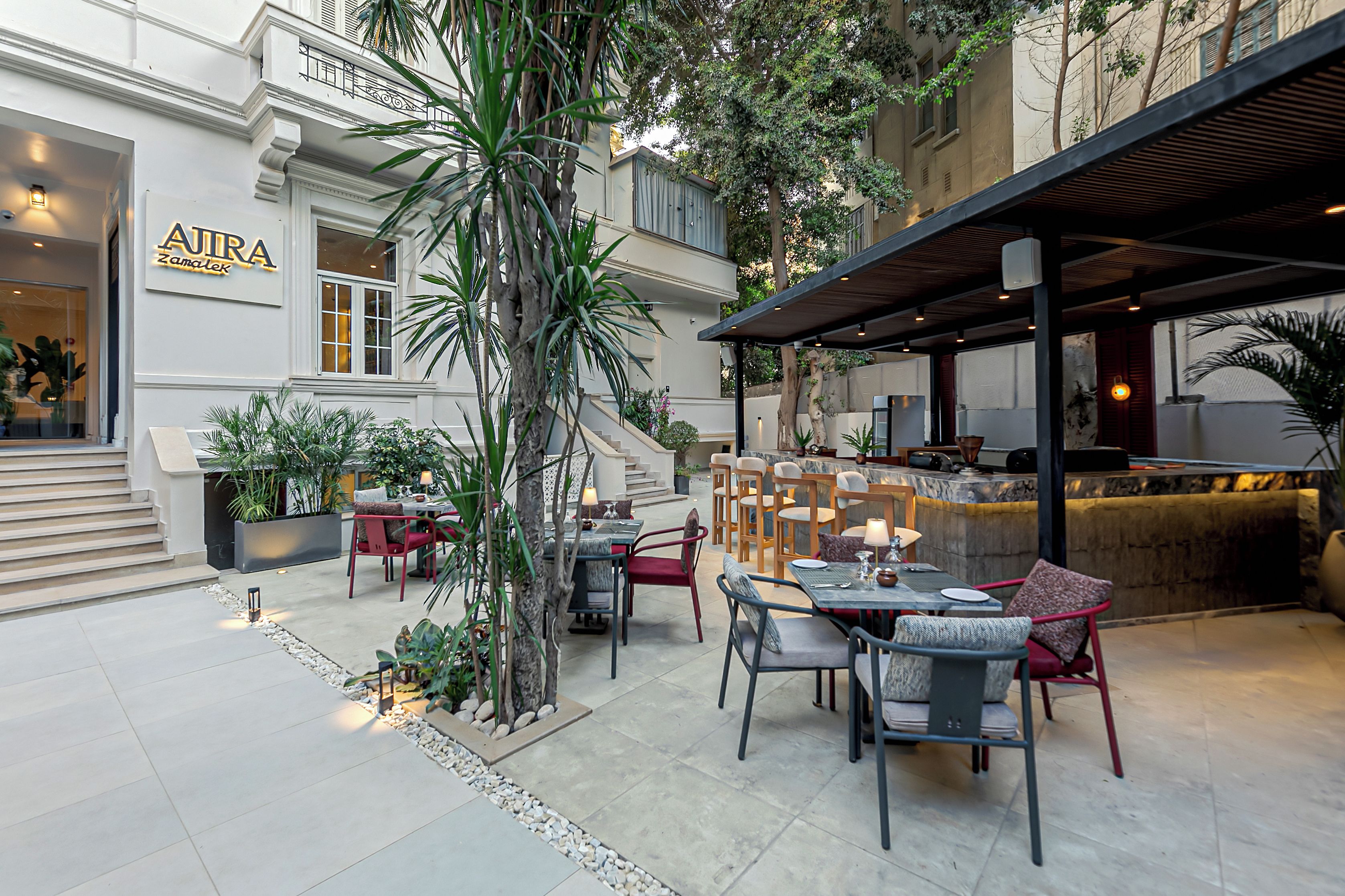 Un área de comedor al aire libre con mesas y sillas, rodeada de plantas y una escalera que lleva a un nivel superior.