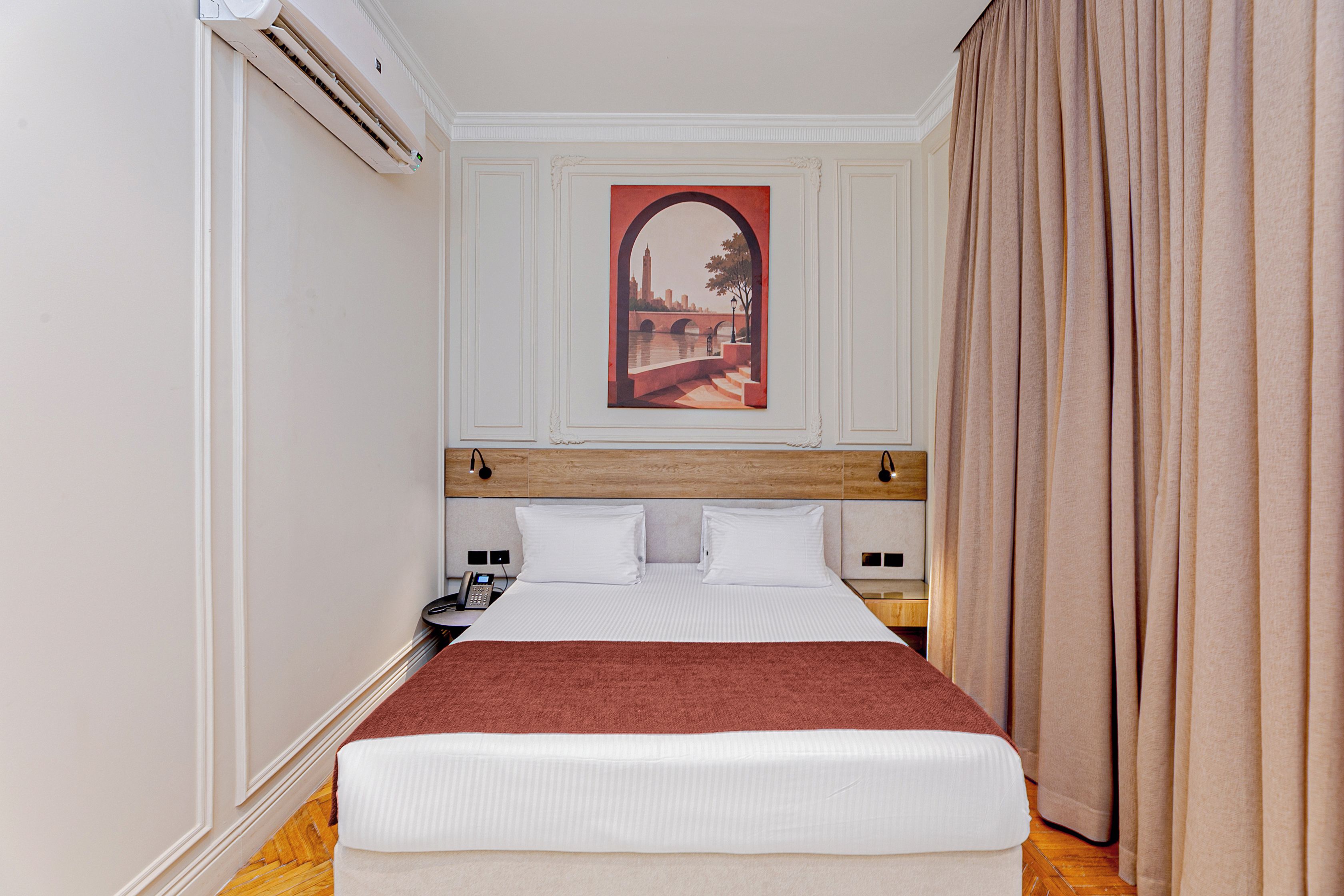 A hotel room with a bed, a painting of a cityscape, a telephone, and a nightstand.