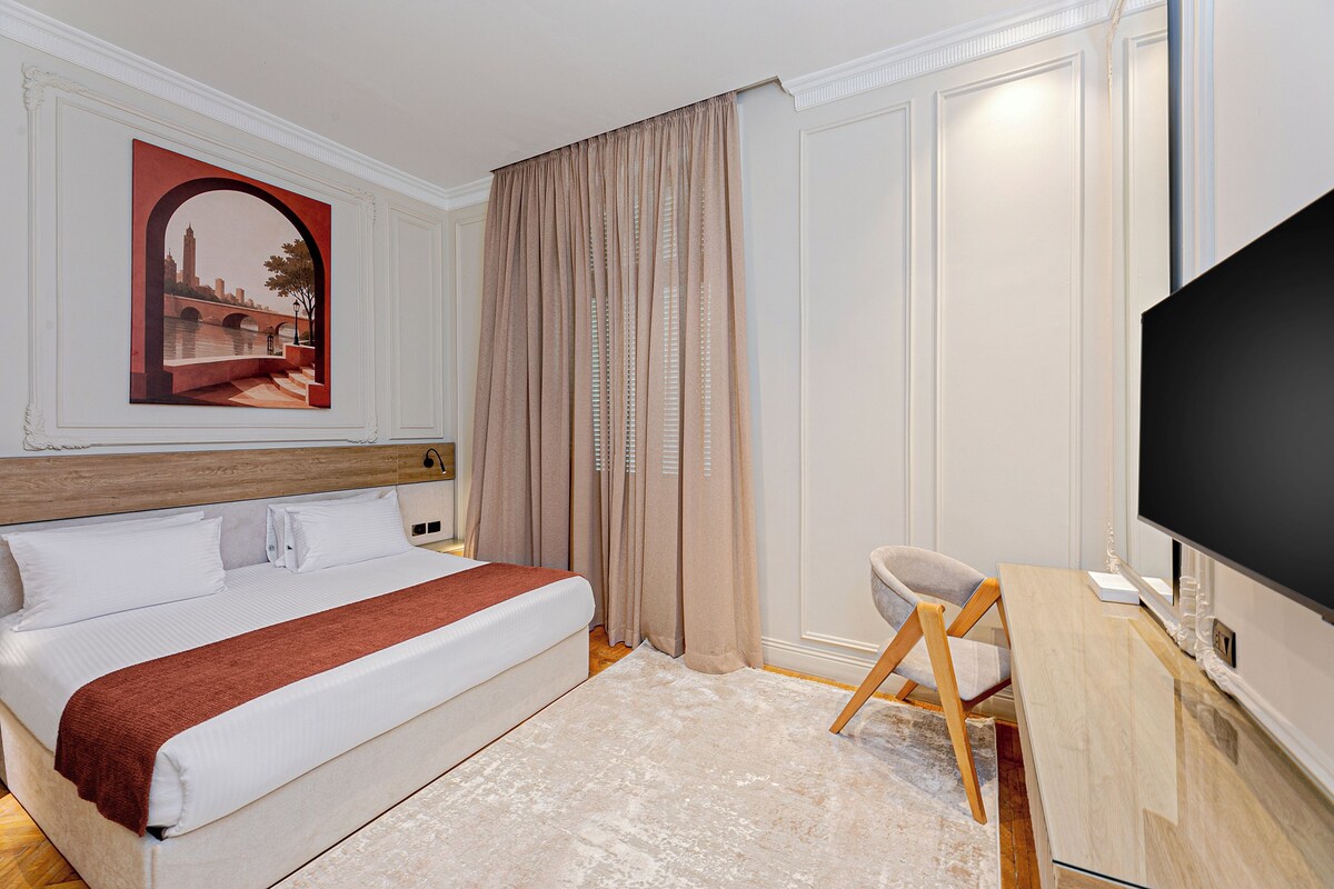 A hotel room with a bed, a desk with a chair, a TV, and a painting on the wall.