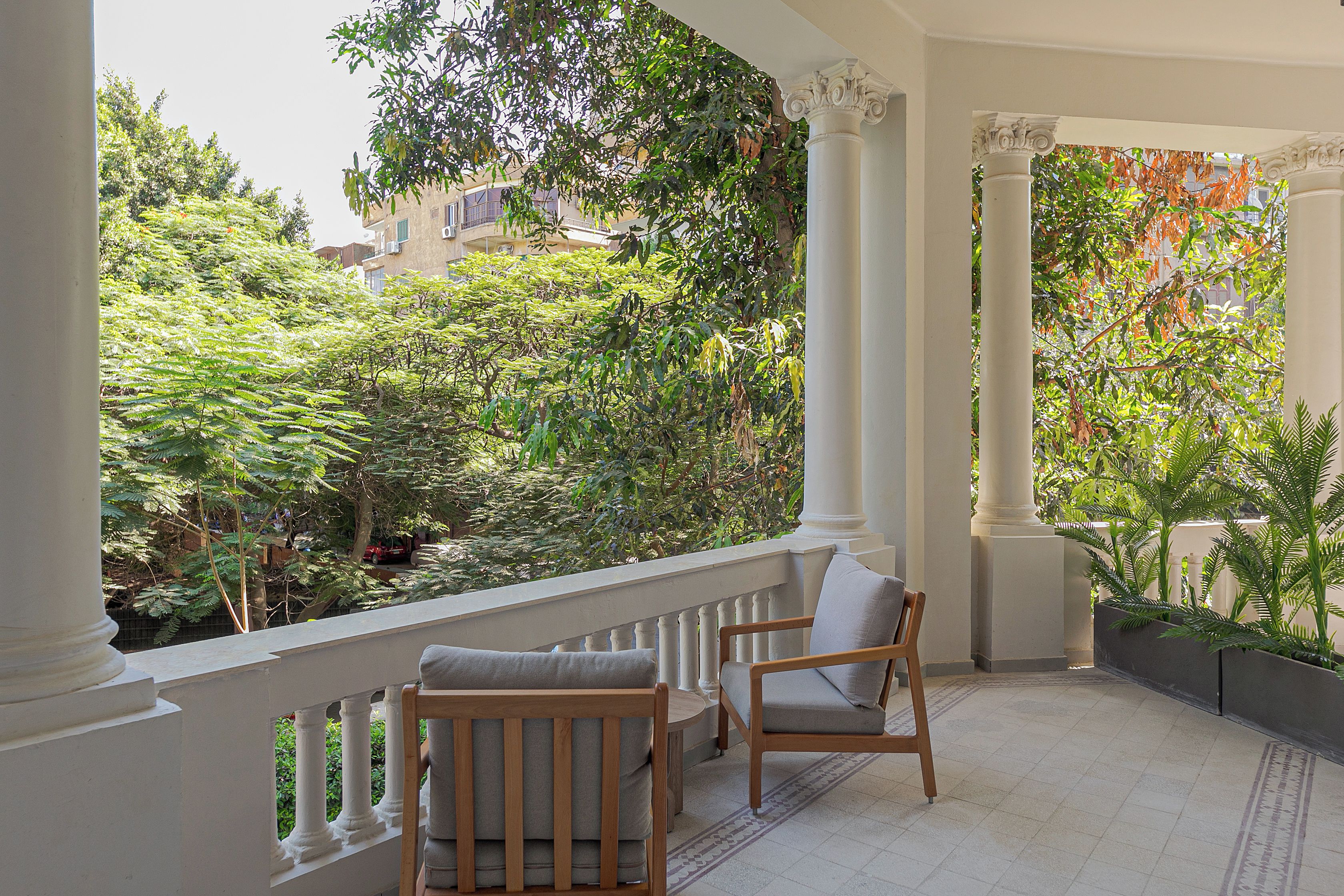 Een balkon met witte zuilen, een houten stoel en uitzicht op een weelderige tuin.