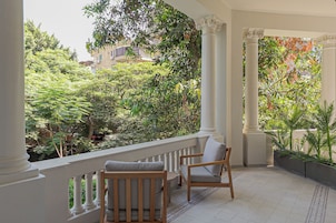 Uma varanda com colunas brancas, uma cadeira de madeira e vista para um jardim exuberante.